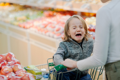 Young boy child crying temper tantrum in shopping cart with mother parent in produce section aisle of supermarket growing pains