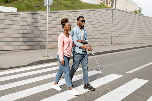 Young black woman assisting visually impaired millennial guy with cane crossing city street. African American blind man with his guide using crosswalk in downtown area. Vision disability concept