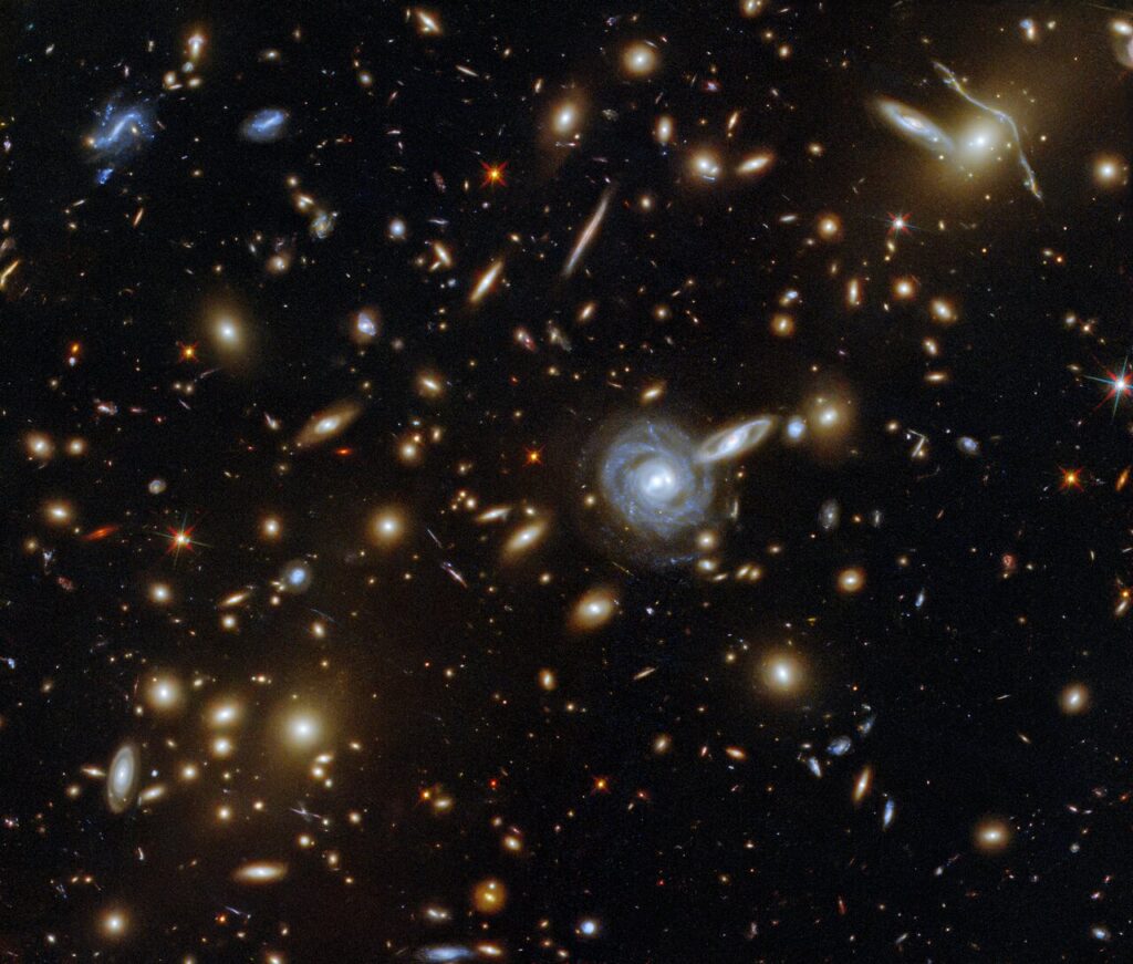 This packed ESA/Hubble Picture of the Week showcases the galaxy cluster ACO S 295, as well as a jostling crowd of background galaxies and foreground stars. Galaxies of all shapes and sizes populate this image, ranging from stately spirals to fuzzy ellipticals. As well as a range of sizes, this galactic menagerie boasts a range of orientations, with spiral galaxies such as the one at the centre of this image appearing almost face on, and some edge-on spiral galaxies visible only as thin slivers of light. The cluster dominates the centre of this image, both visually and physically. The huge mass of the galaxy cluster has gravitationally lensed the background galaxies, distorting and smearing their shapes. As well as providing astronomers with a natural magnifying glass with which to study distant galaxies, gravitational lensing has subtly framed the centre of this image, producing a visually striking scene. Links  Video of a Menagerie of Galaxies 