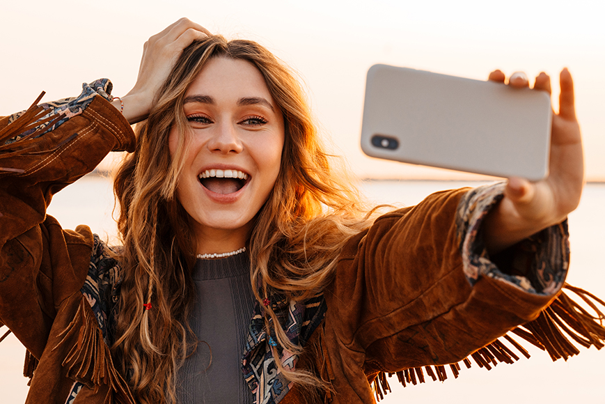 Image of a excited happy young woman walking outdoors at the beach and taking a selfie by smartphone