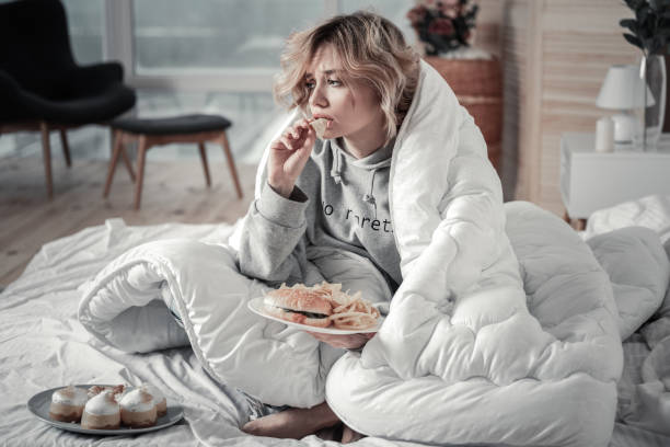Burger and fries. Sad and lonely blonde-haired young woman eating burger and French fries in the bed
